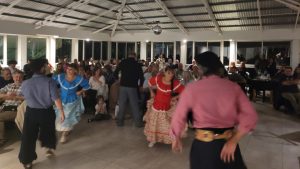 En la imágen se muestran dos parejas de bailarines con vestimentas típicas tradicionalistas en el medio de un salón con gente.